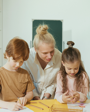Eine Frau sitzt mit zwei Kindern an einem Tisch und arbeitet mit ihnen an Papier und Stiften